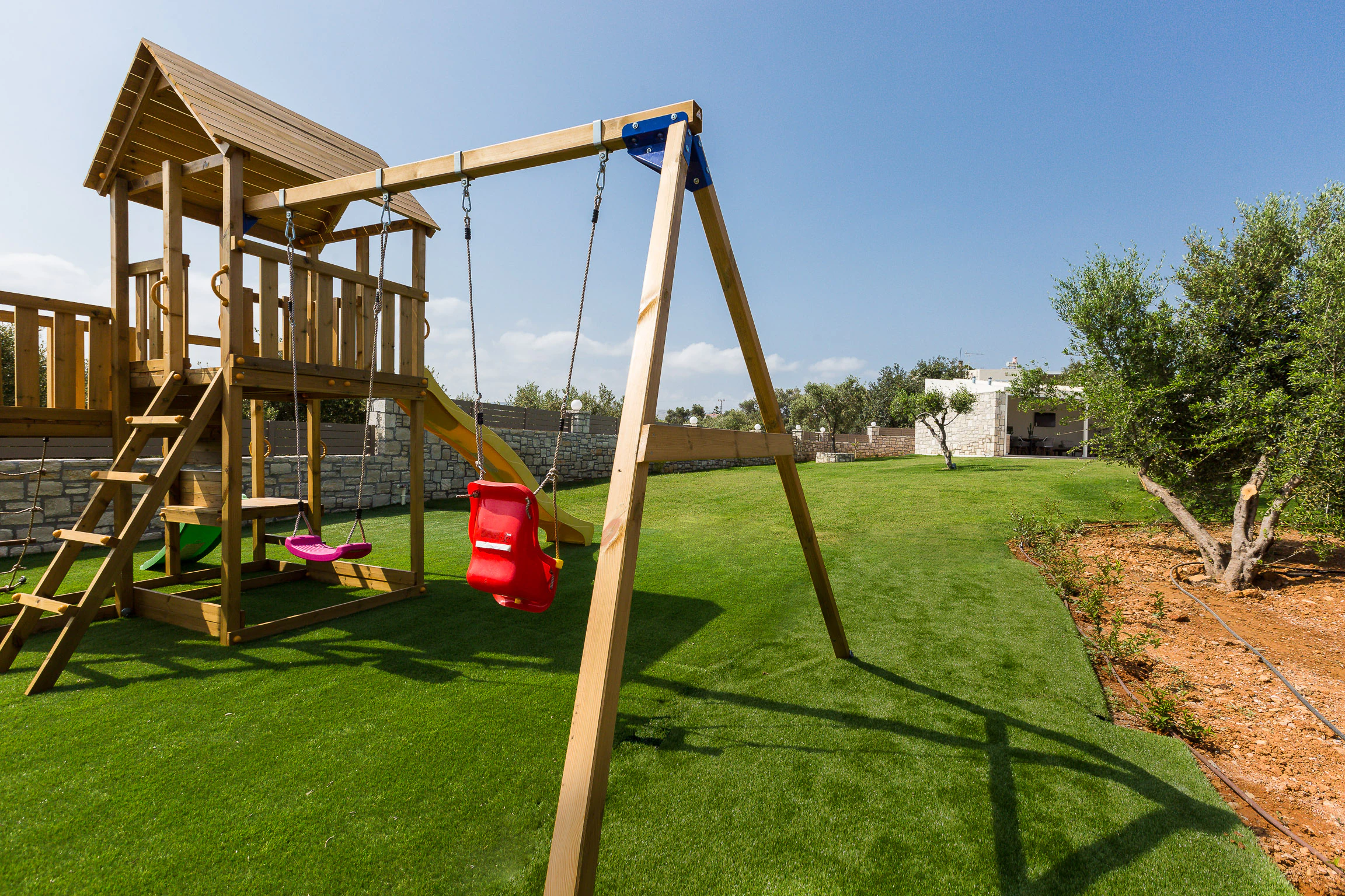 A wooden climbing frame with a slide and swings on the lawn beside olive trees