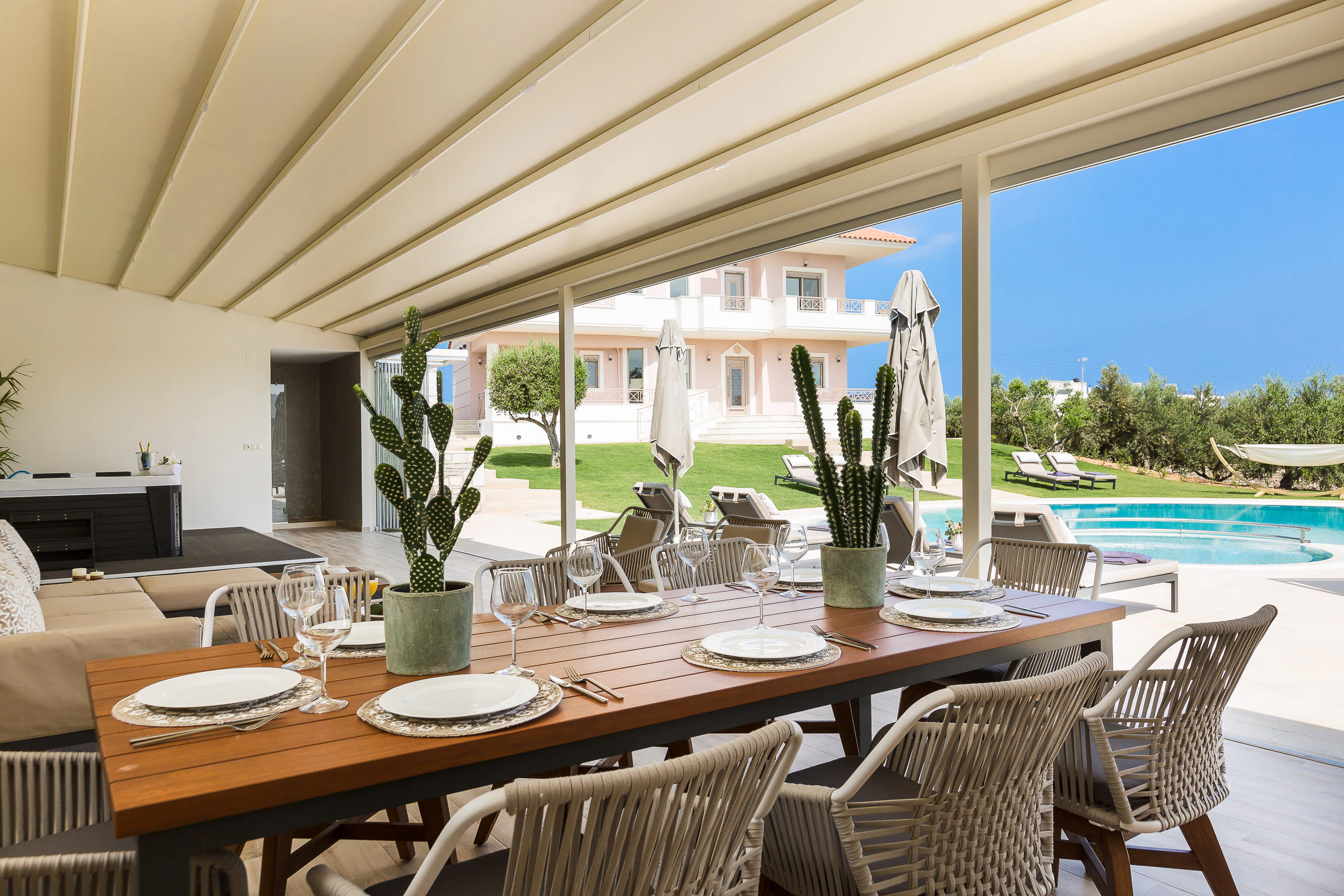 A long outdoor dining table laid for eight under the covered kitchen, open to the pool and lawn